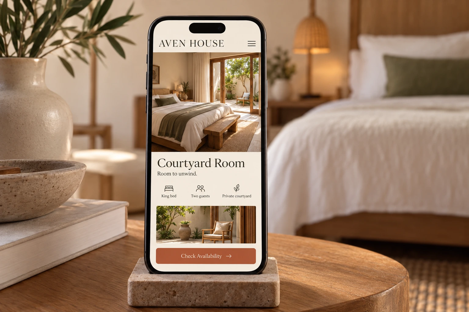 A phone on a stone stand shows the Aven House Courtyard Room page, with a bedroom photo, “Room to unwind.” and a terracotta “Check Availability” button.