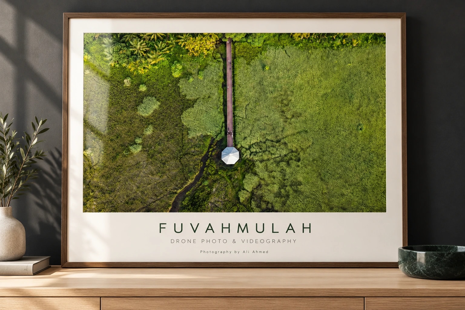 A framed Fuvahmulah print of a boardwalk cutting through green wetland from above, with “DRONE PHOTO & VIDEOGRAPHY” beneath, on a wooden sideboard against a dark wall.
