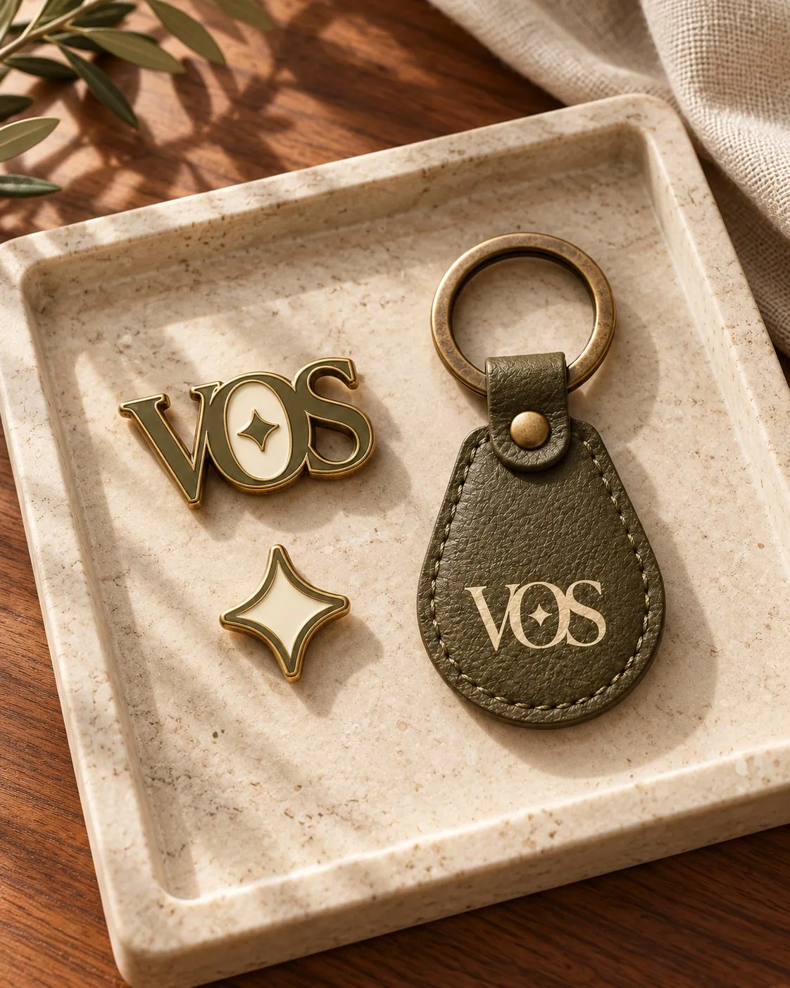 A VOS enamel pin, a star pin and an olive leather key ring with a brass ring, on a stone tray.